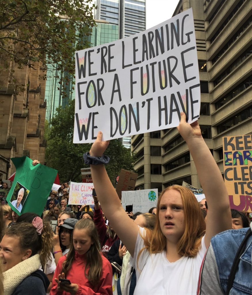 A teenage girl holding a hand-written banner amongst a crowd of people at Sydney Town Hall protesting against inaction on climate change. The banner reads 'We're leaning for a future we don't have'.